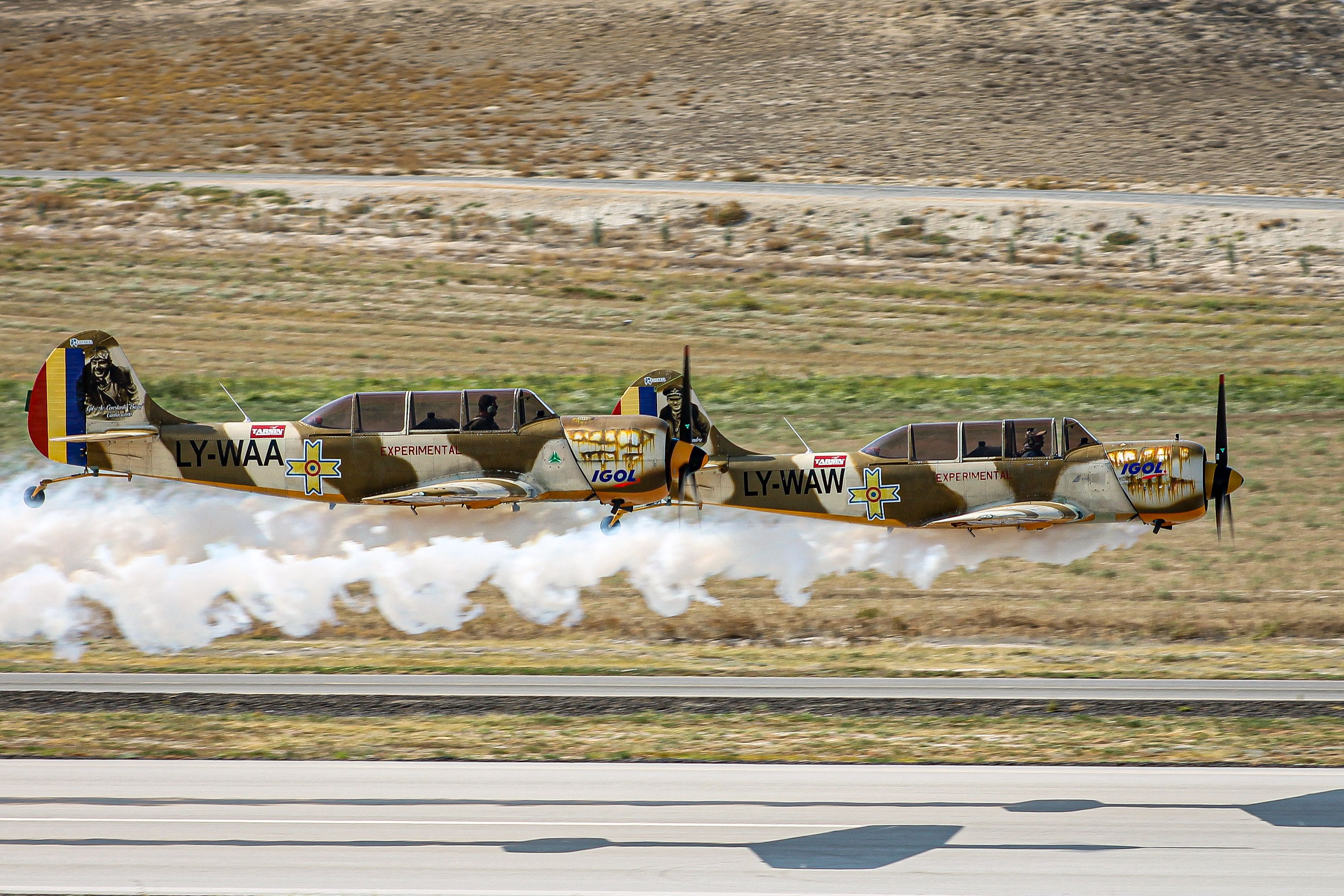 Two Yakovlev Yak-52 planes in synchronized flight with smoke trails over a field.