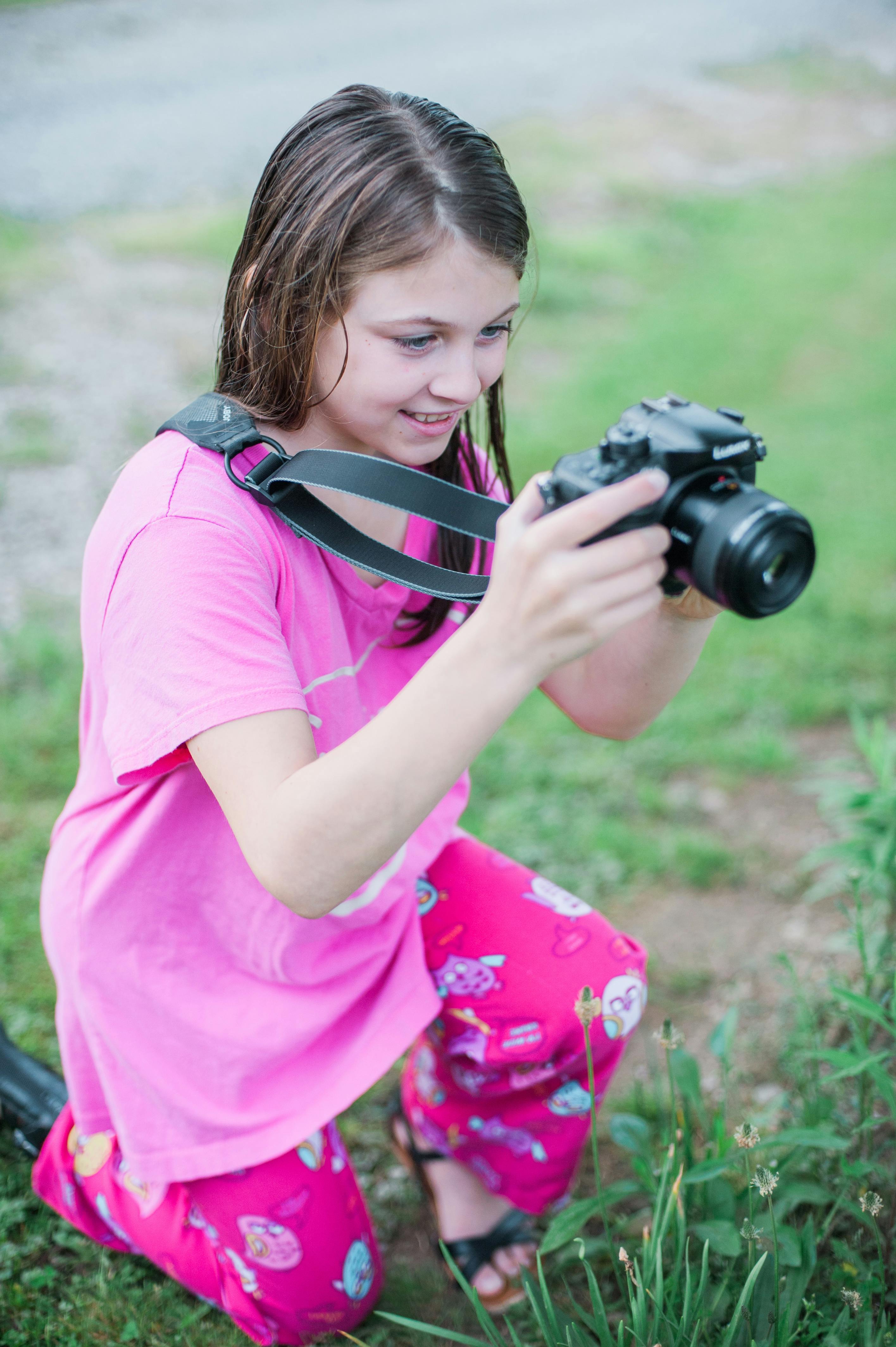 黒のデジタル一眼レフカメラを保持しているピンクの丸首のシャツを着た女の子の浅い焦点の写真 無料の写真素材