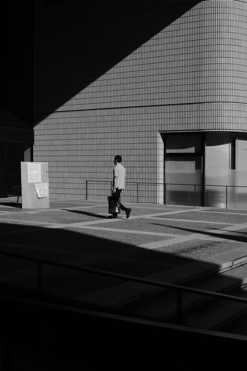 Free Black and white photo of a man walking in an urban setting, showcasing architectural shadows. Stock Photo