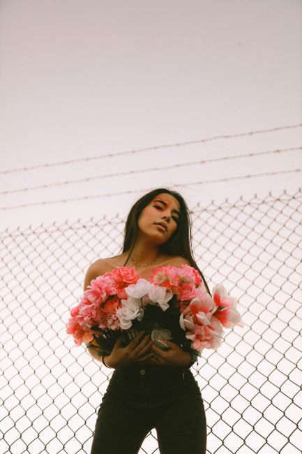 Young woman with long hair holding flowers, standing in front of a chain-link fence under a clear sky.