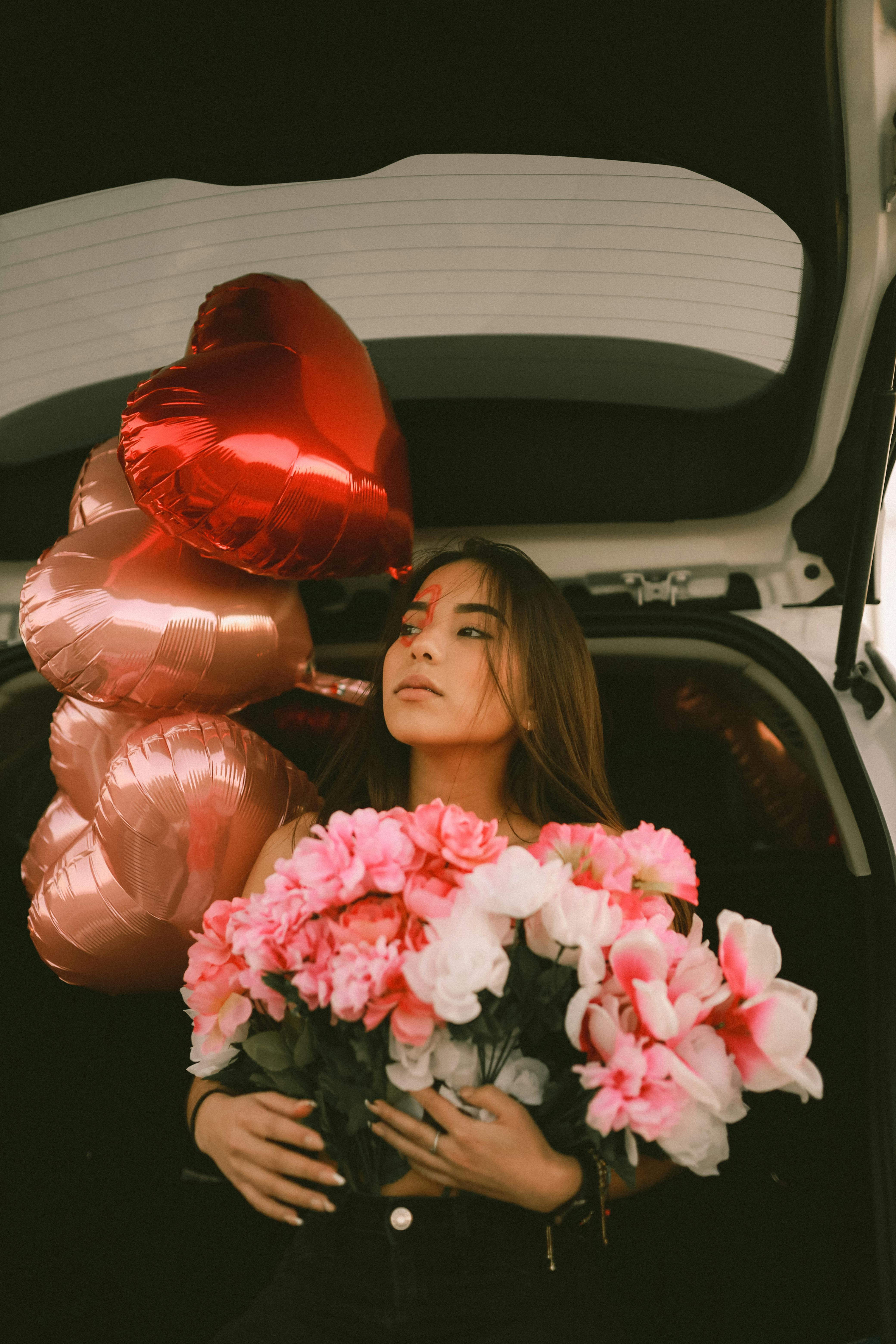 A young woman holding a floral bouquet with heart balloons in a car trunk, ideal for Valentine's Day themes.