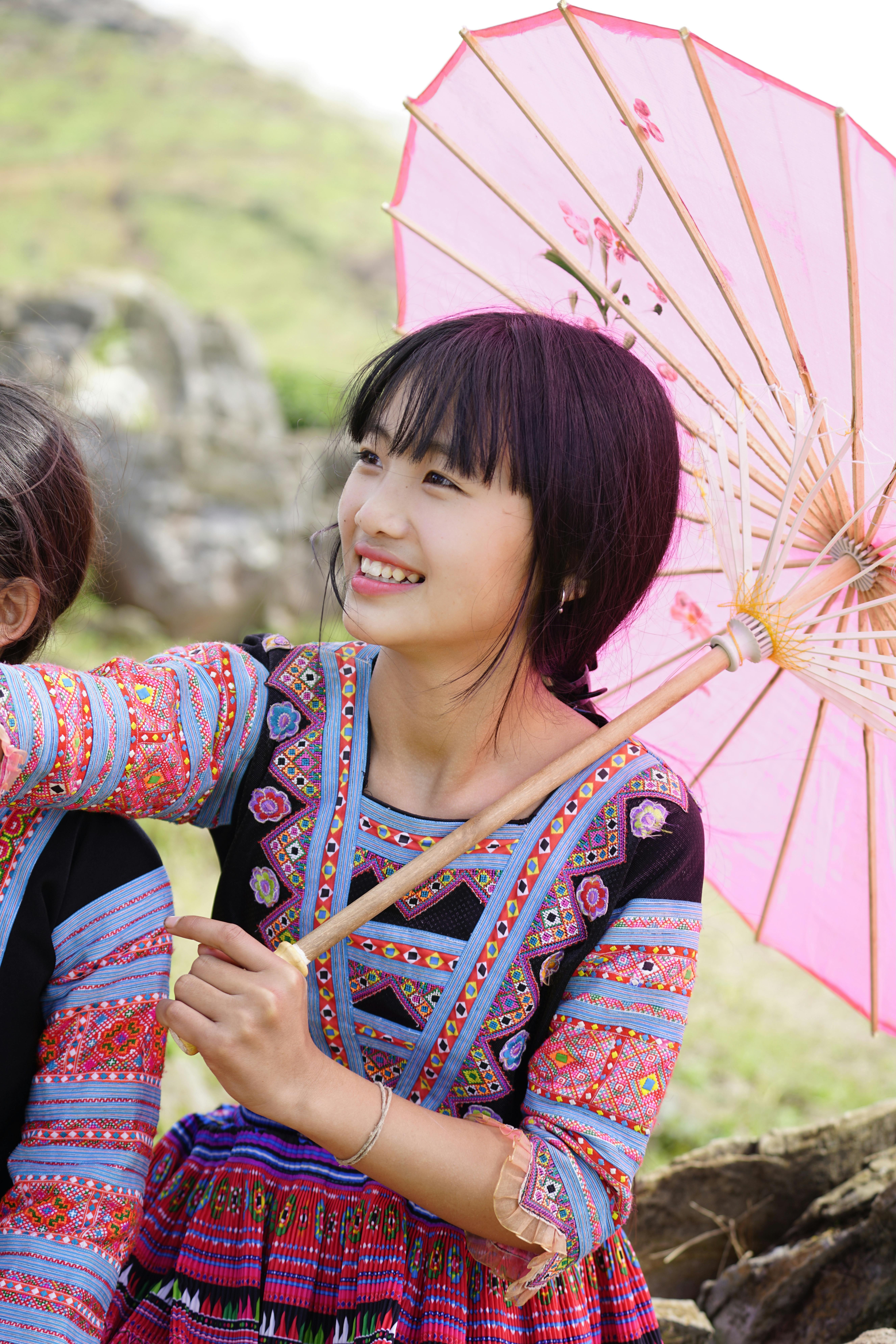 A young woman joyfully smiles while holding a pink parasol, dressed in vibrant traditional clothing outdoors.