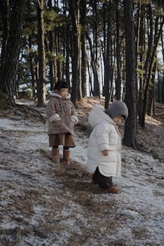 Two children exploring a snowy forest in winter, wearing warm jackets.