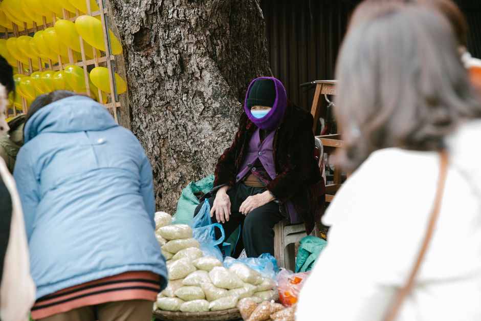 Dark-skinned Nigerian market trader, a middle-aged woman wearing a yellow Ankara wrapper and gele headtie, sitting behind large bags of white and yellow garri displayed in open basins at a busy Bodija Market stall in Ibadan, customers browsing in the background, warm afternoon sunlight, photorealistic