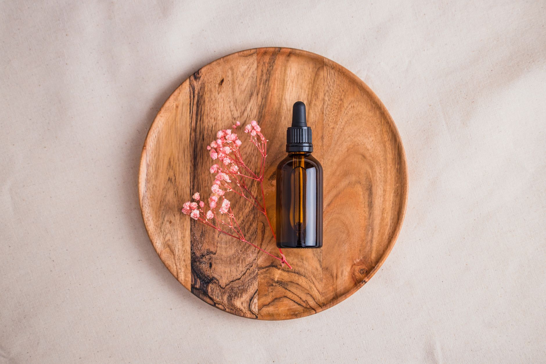 Top view of a brown glass bottle with dropper on wooden plate, accompanied by pink flowers, showcasing natural cosmetics.