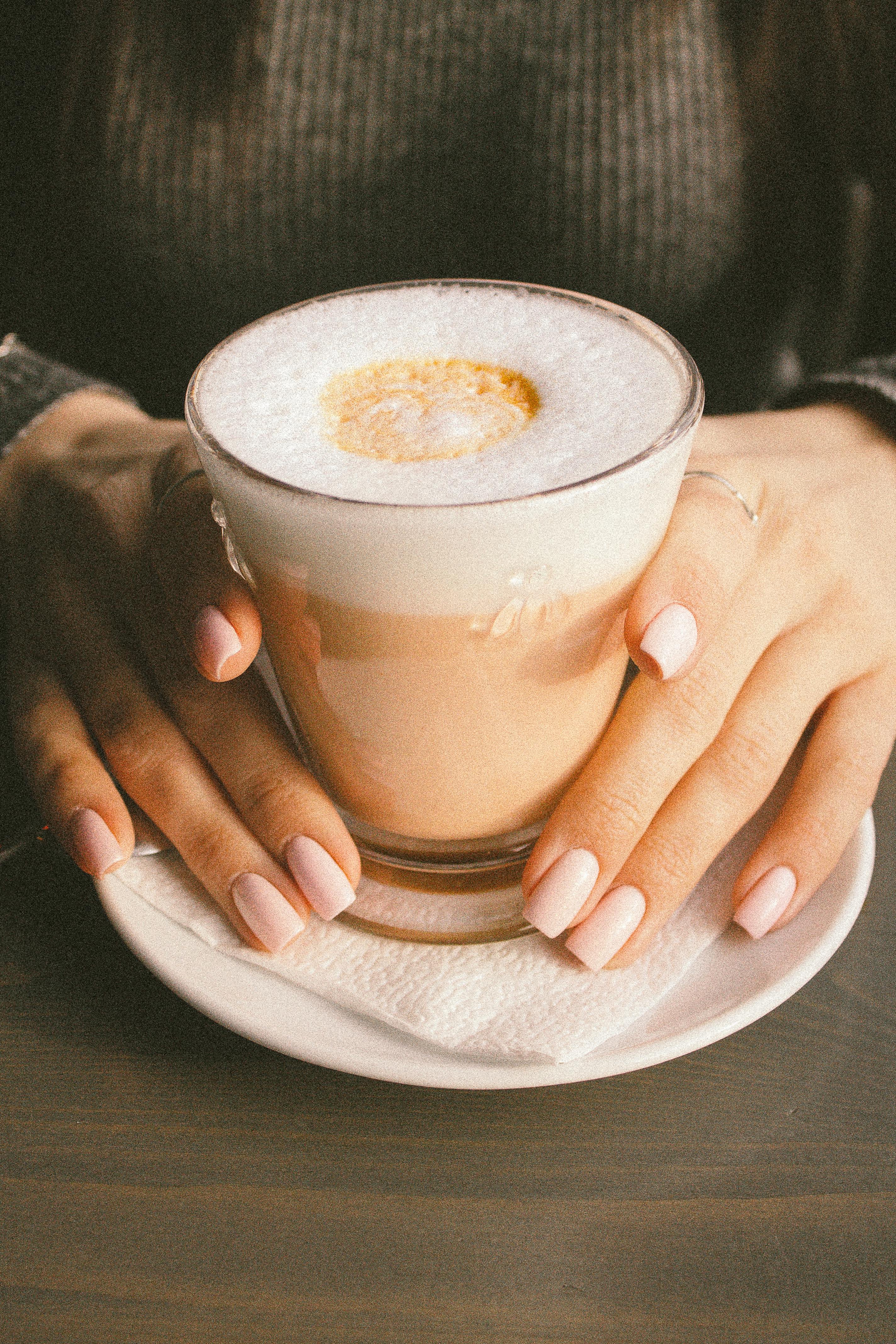 A warm and inviting photo of a latte with foam held by manicured hands in a cafe.