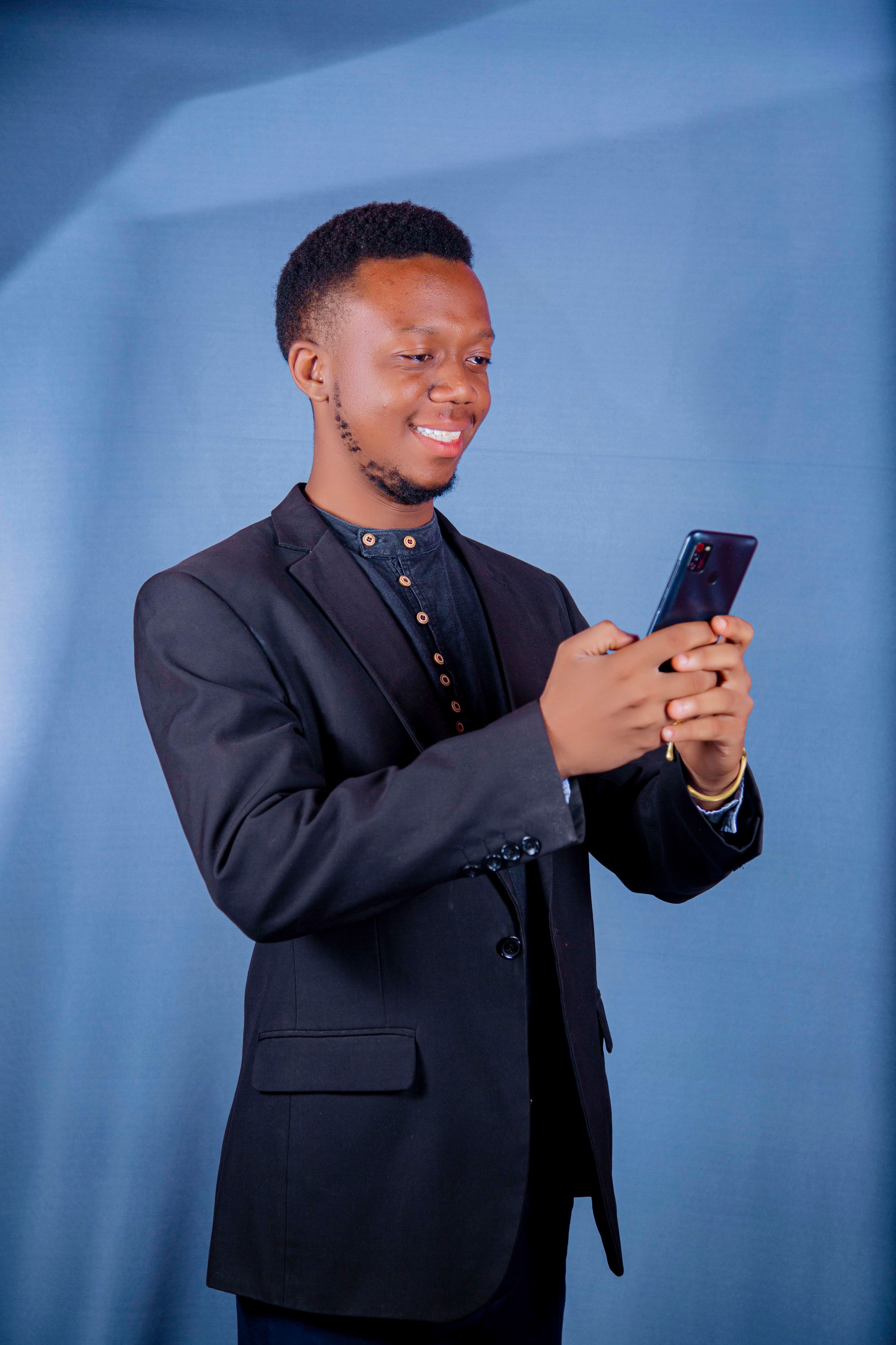 An adult African American man smiles while using a smartphone indoors, dressed in a business blazer.