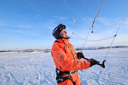 A man wearing orange warm clothing and safety helmet paragliding over a snowy field on a sunny winter day.