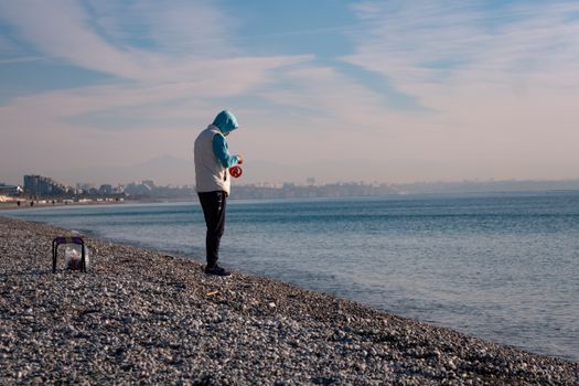 A man in a hoodie fishing alone on a rocky beach, overlooking a calm sea under a bright sky.