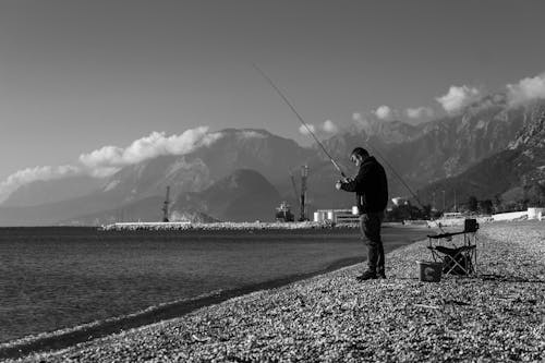 A Grayscale Photo of a Man in Black Jacket Standing on Shore while Holding a Fishing Rod