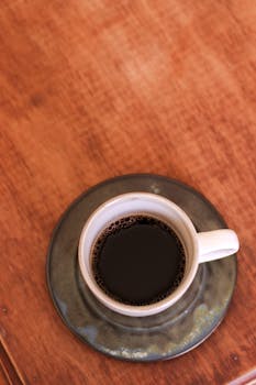 Aerial shot of black coffee in a ceramic cup with saucer on a wooden table, emphasizing elegant simplicity.