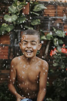 Smiling child enjoying the rain outdoors, surrounded by lush plants, perfect for lifestyle and happiness themes.
