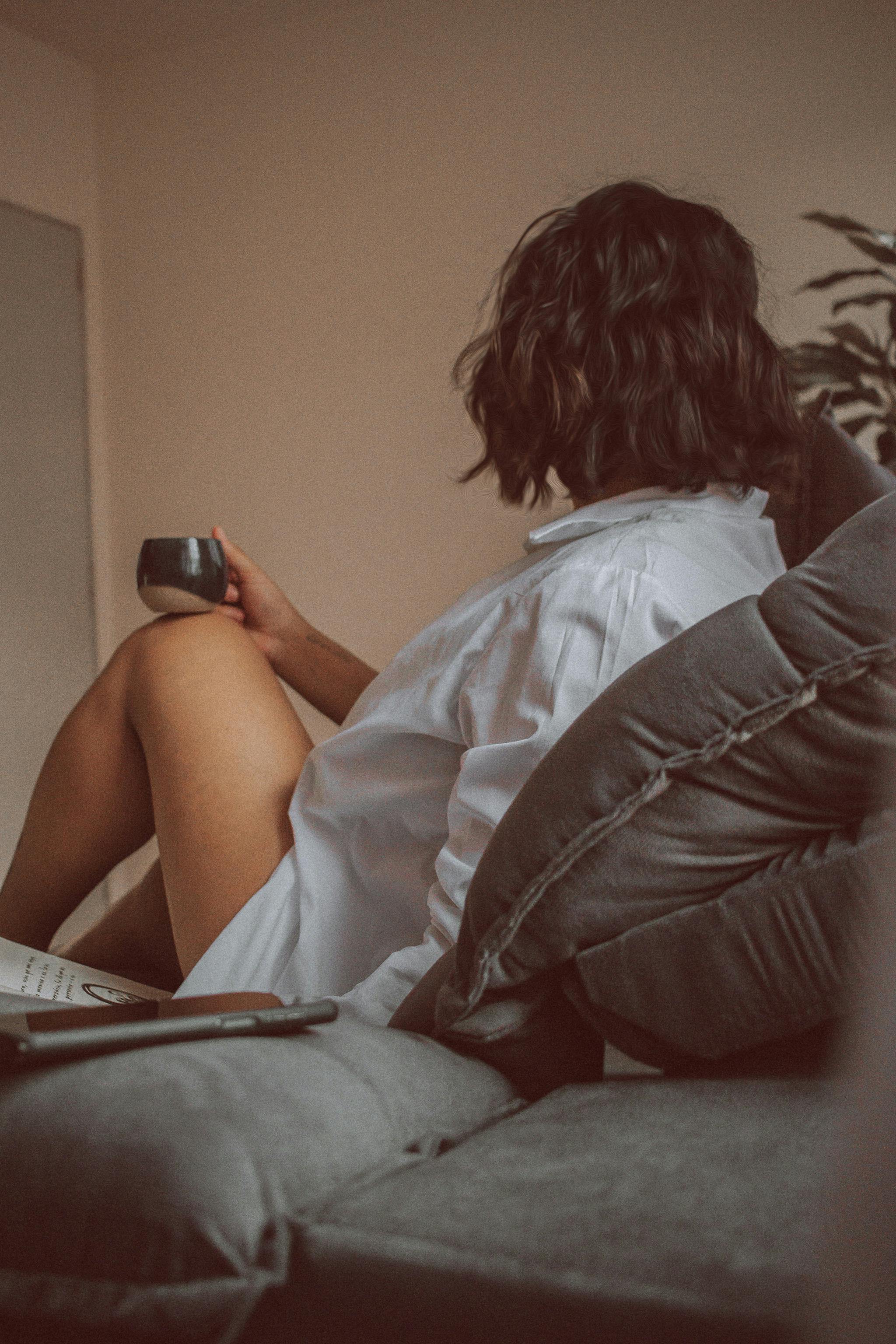 Woman in casual white shirt holding a cup while sitting in a cozy indoor space, capturing a calm and relaxed moment.