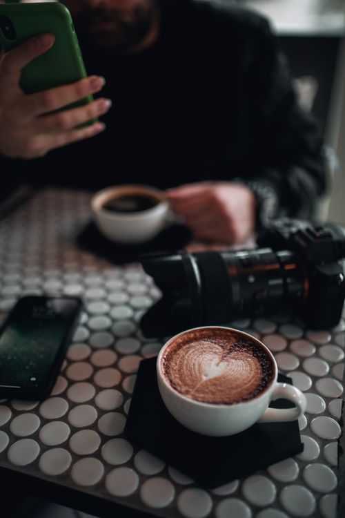 Free Close-up of a latte with intricate foam art on a stylish café table with a camera in view. Stock Photo