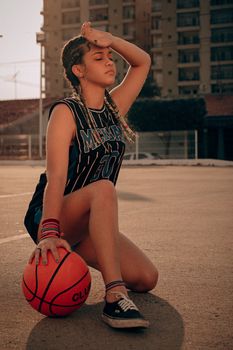 A young woman with braided hair rests on an outdoor court, holding a basketball.