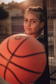 Confident young woman with braided hair holds a basketball outdoors in warm lighting.