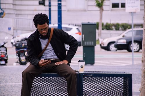 Free Man sitting on a street bench in San Francisco, checking his phone with a coffee cup nearby. Stock Photo
