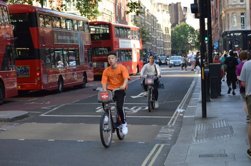 Free Young men cycling alongside iconic red buses in a vibrant London street scene. Stock Photo