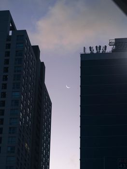 A crescent moon visible in the evening sky between two city skyscrapers, creating a serene urban silhouette.