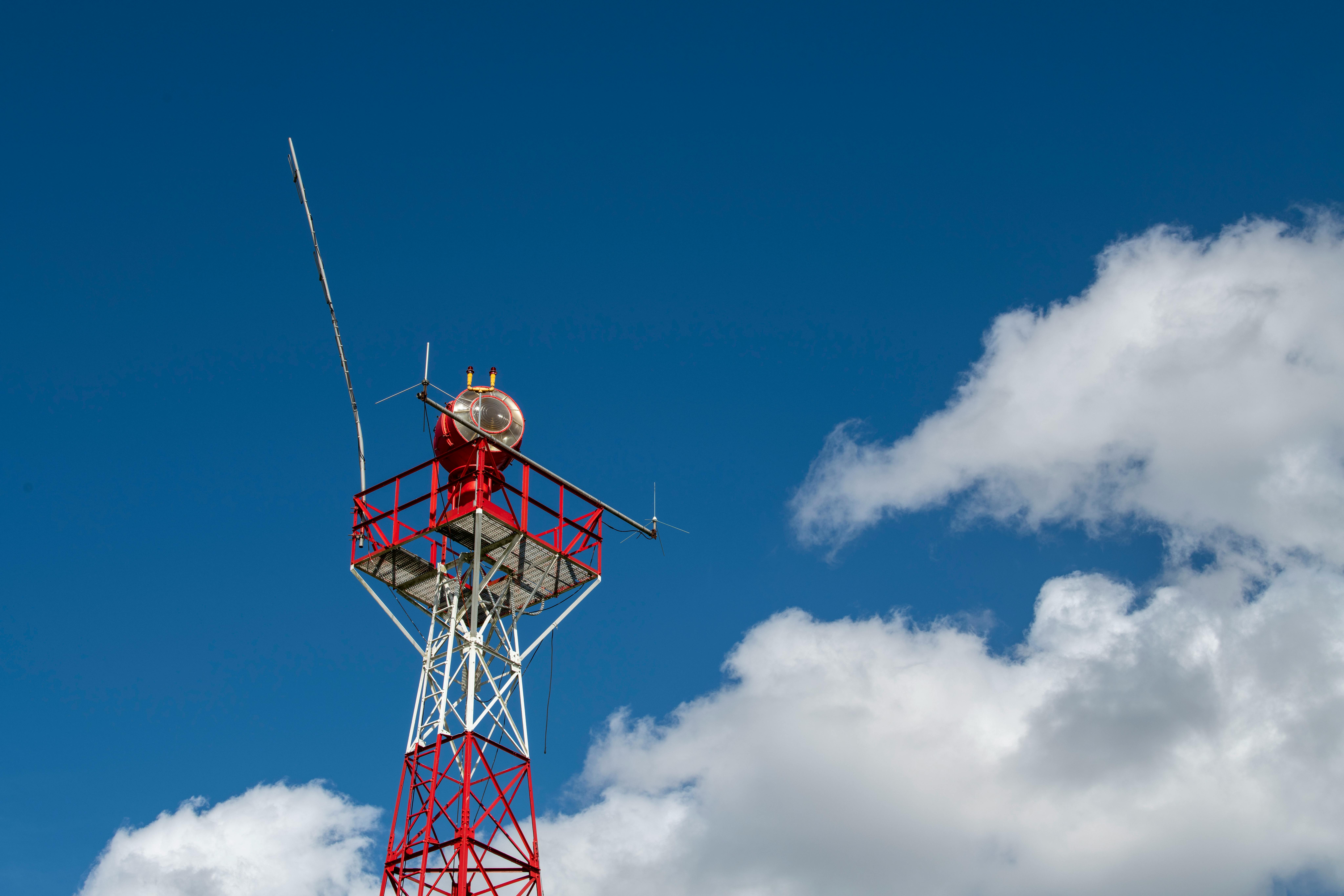 Low-angle shot of a red and white communication tower reaching into a bright blue sky with fluffy cl