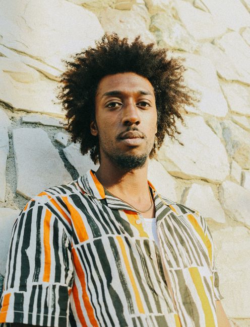 Young man with curly hair in a striped shirt standing against a stone wall in sunlight.