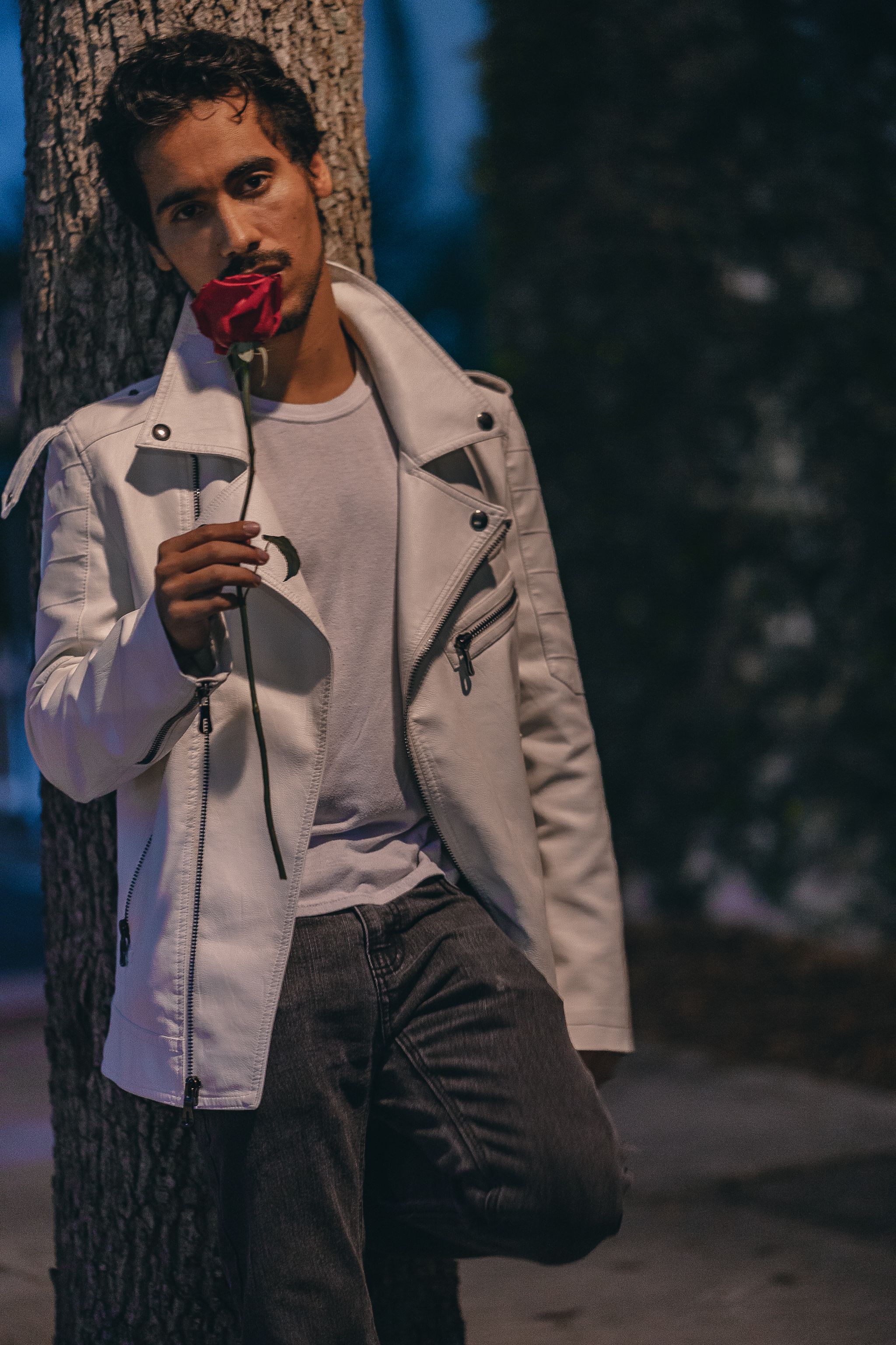 A stylish young man in a white jacket poses with a red rose at night outdoors.