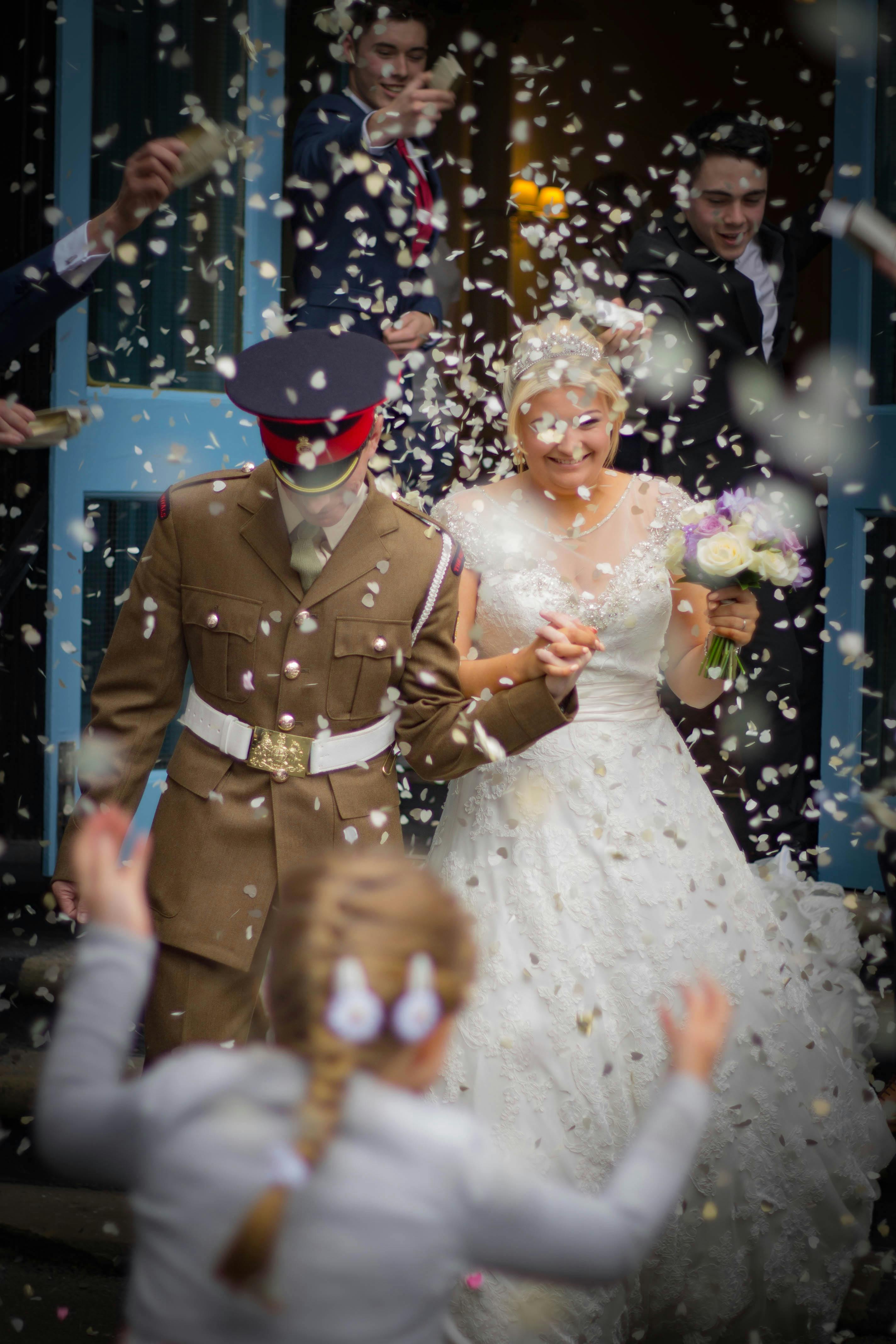 A cheerful wedding exit adorned with confetti, featuring a bride and groom in military attire.