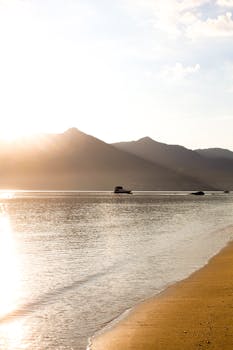 Peaceful beach scene at sunrise with boat silhouette and mountains in the background.