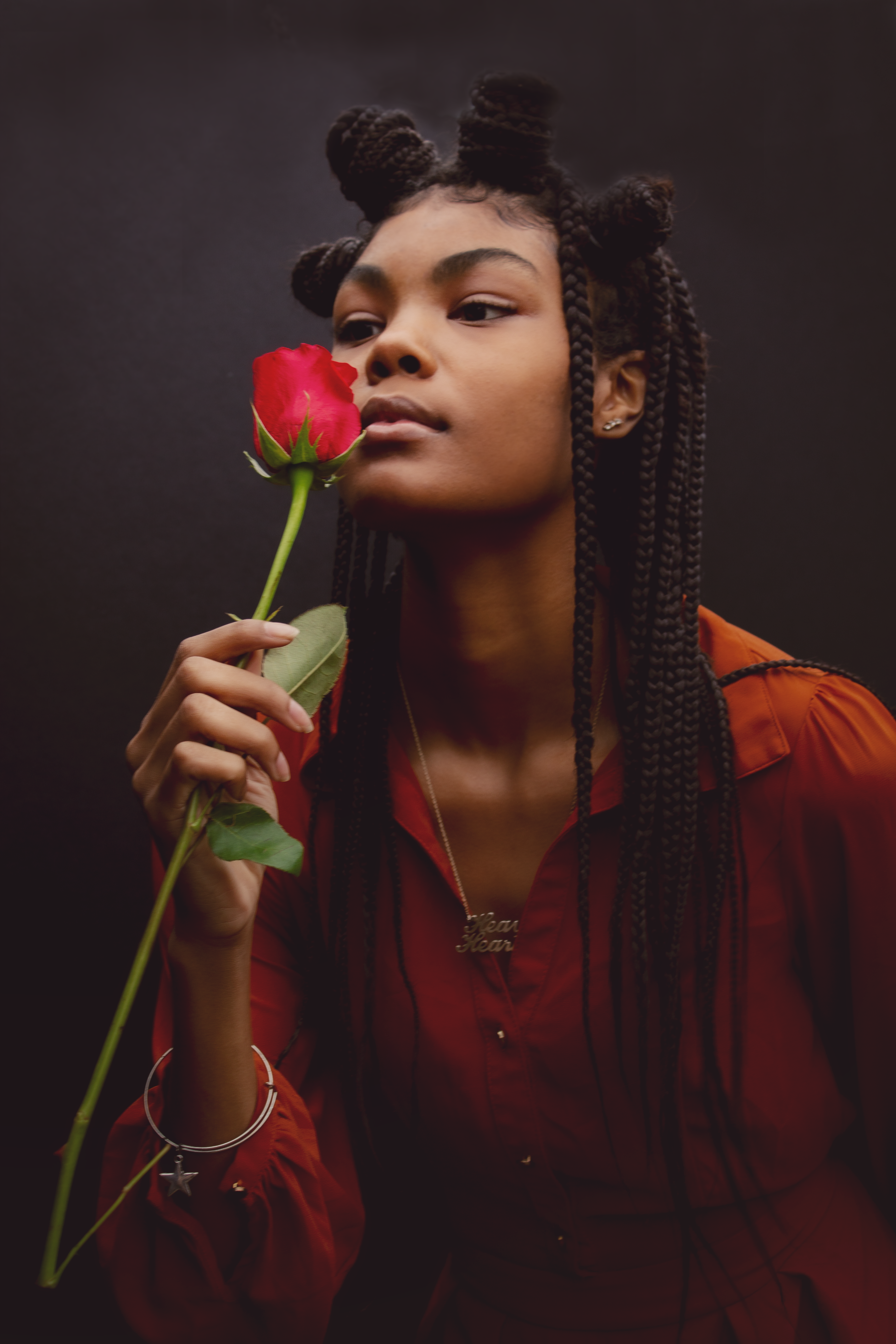 Portrait of a young black woman holding and smelling a red rose, wearing an orange blouse against a dark background.
