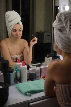 Caucasian woman with towel on head applying makeup in mirror reflection.
