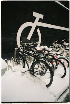 A row of bicycles covered in snow parked against a wall with a large cycle symbol.
