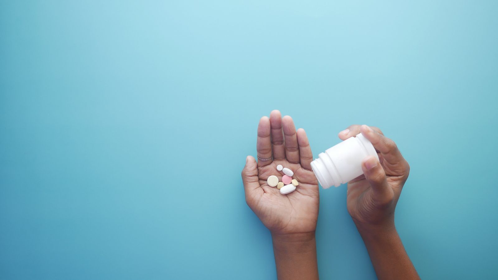 Hands pouring pills from a bottle on a blue background, representing healthcare and medication.