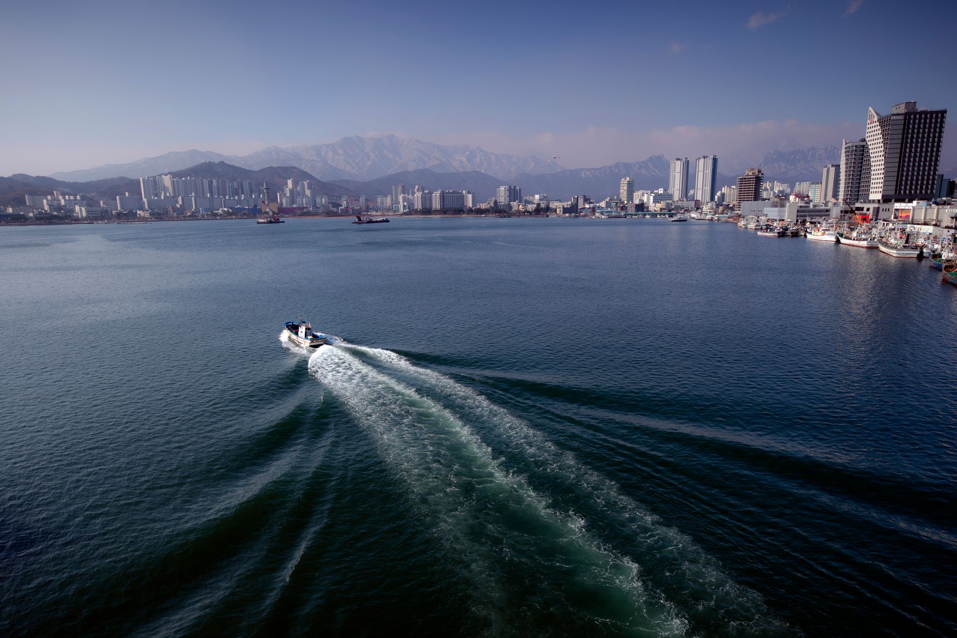Aerial shot of Sokcho's urban waterfront with a motorboat navigating the vast sea under clear skies.