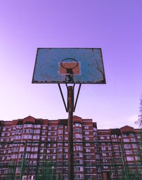 An old basketball hoop stands tall against a cityscape backdrop with a vibrant purple sky.