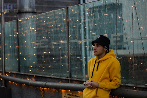 A Man in Yellow Sweater Leaning Near the Glass Wall with String Lights