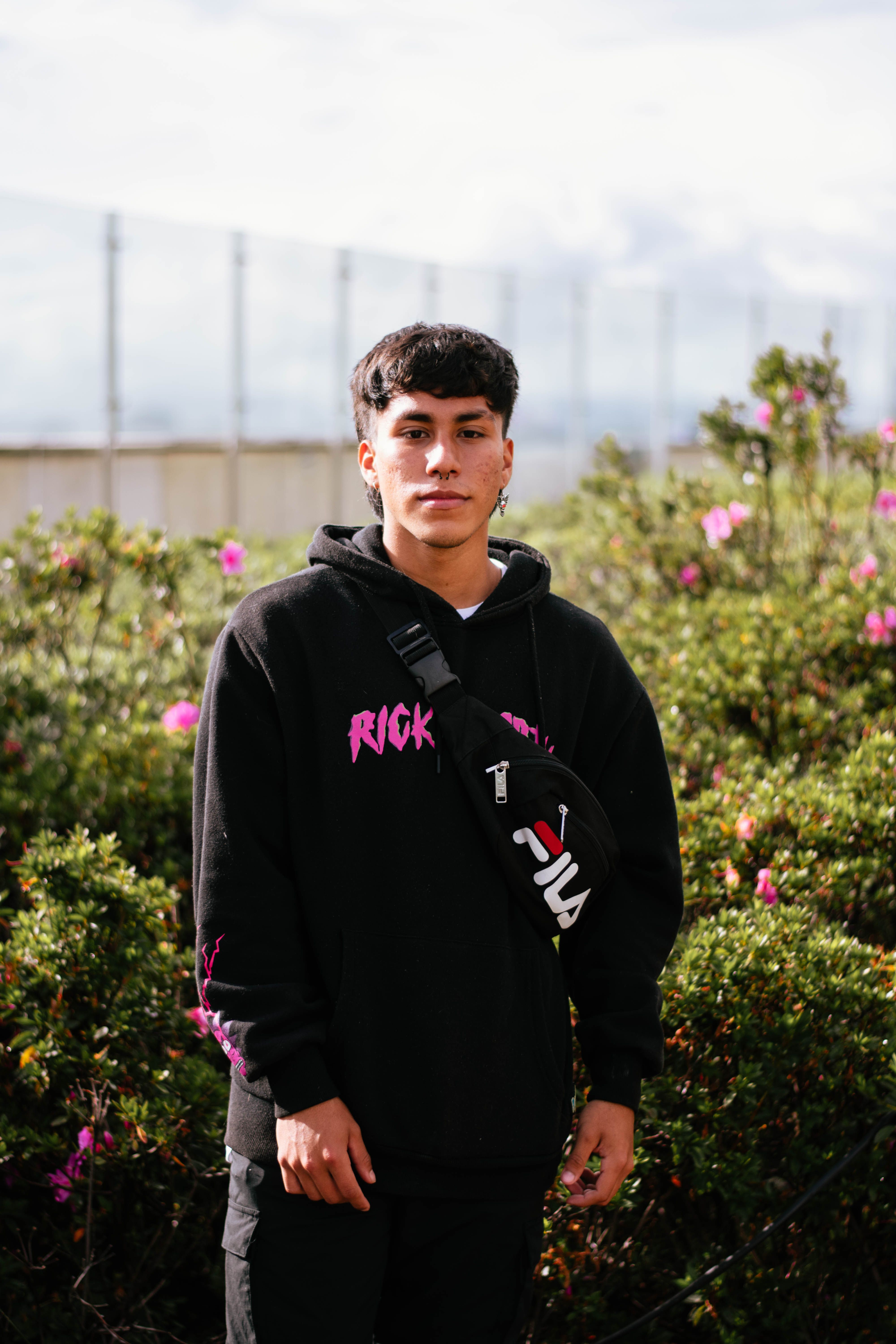 Portrait of a young man wearing a black hoodie standing amidst vibrant blooming shrubs outdoors.