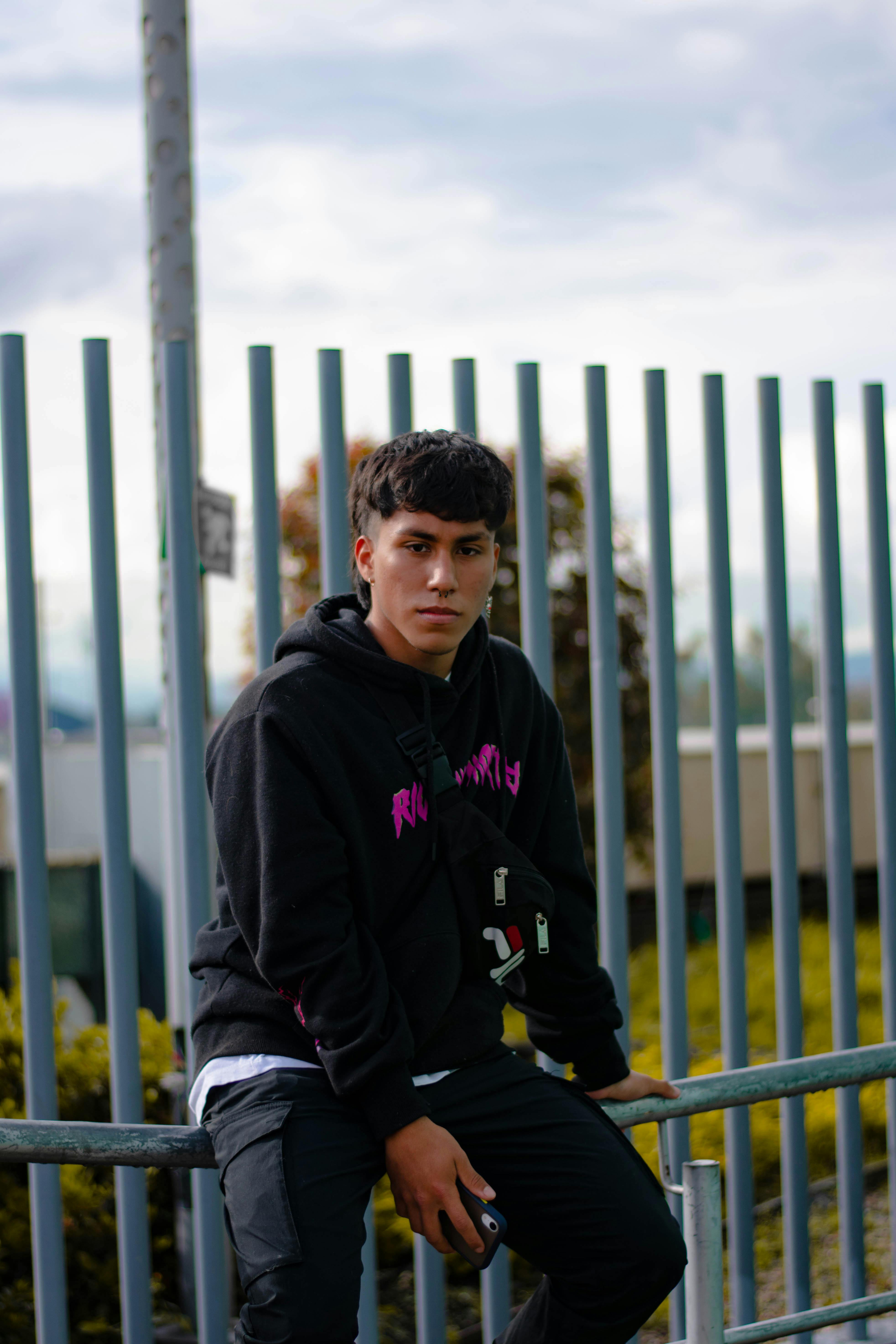 Serious young man with nose piercing sitting on a metal railing holding a smartphone.
