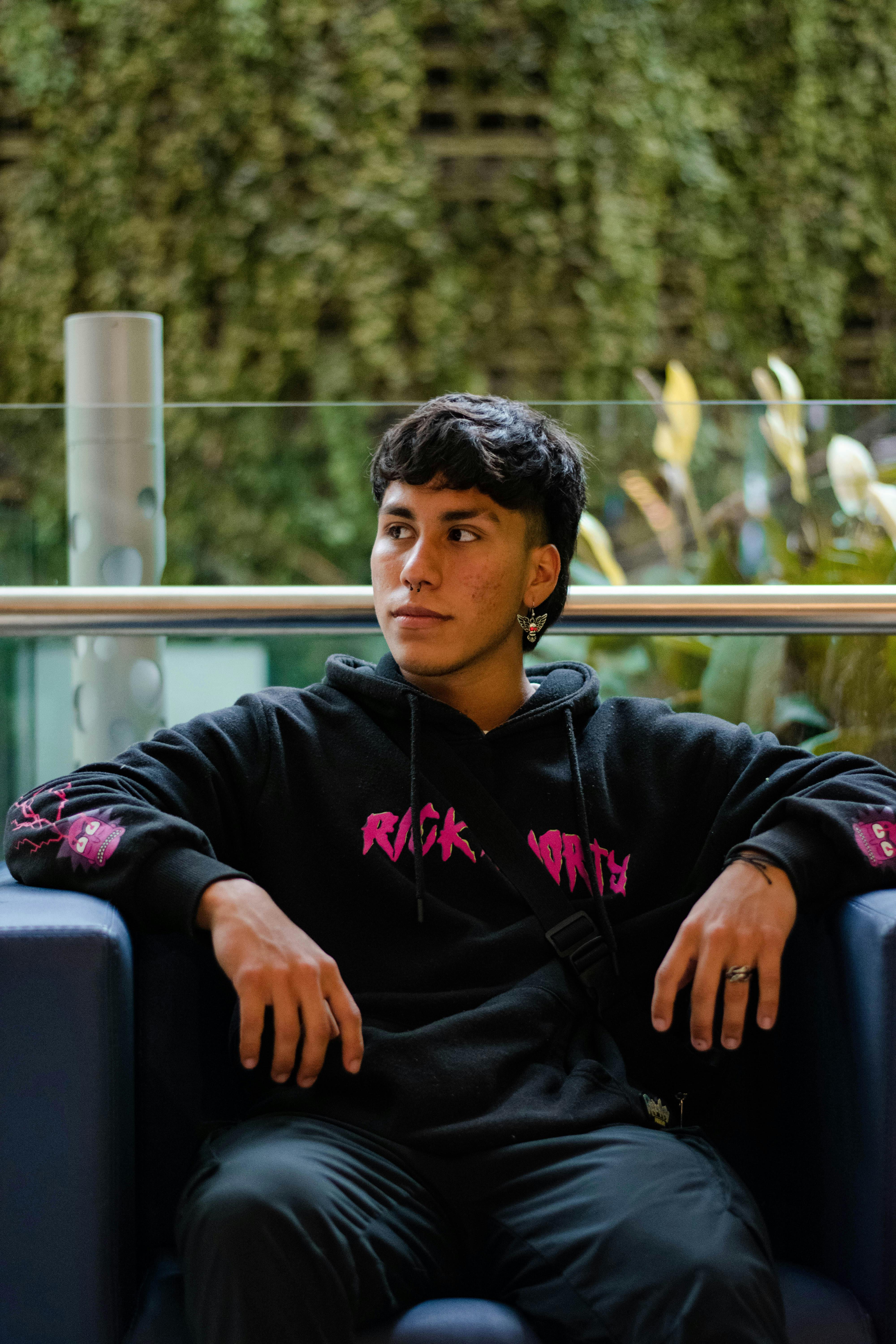 Young man in a black hoodie sitting indoors on a modern armchair.