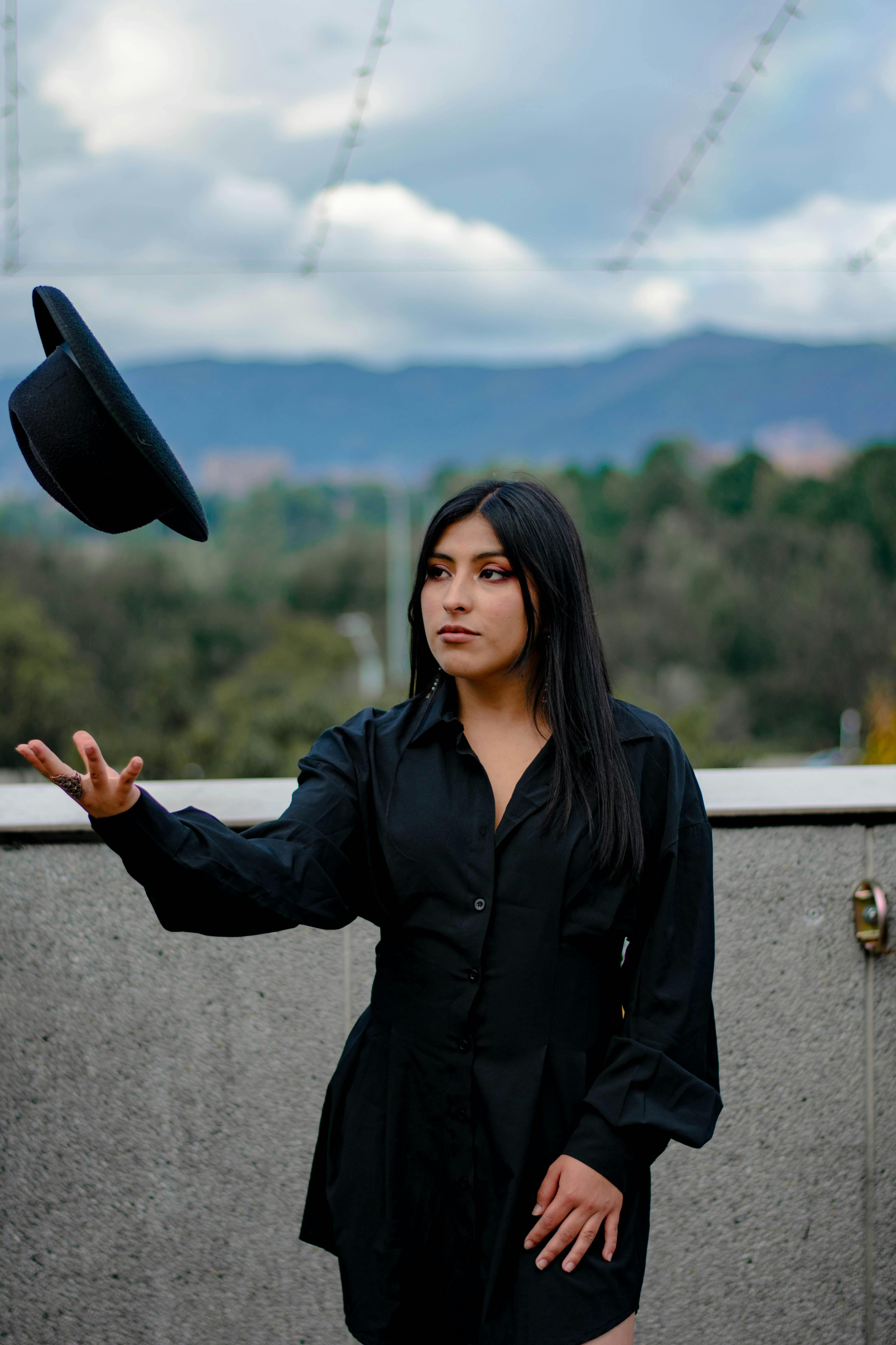 Portrait of a woman in black throwing a hat outdoors with a mountainous backdrop.