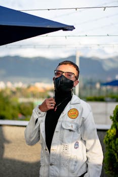 Stylish man in denim jacket and face mask standing outdoors with mountains in the background.