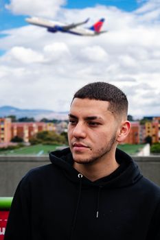 Portrait of a man in a black hoodie outdoors with a flying airplane in the background.
