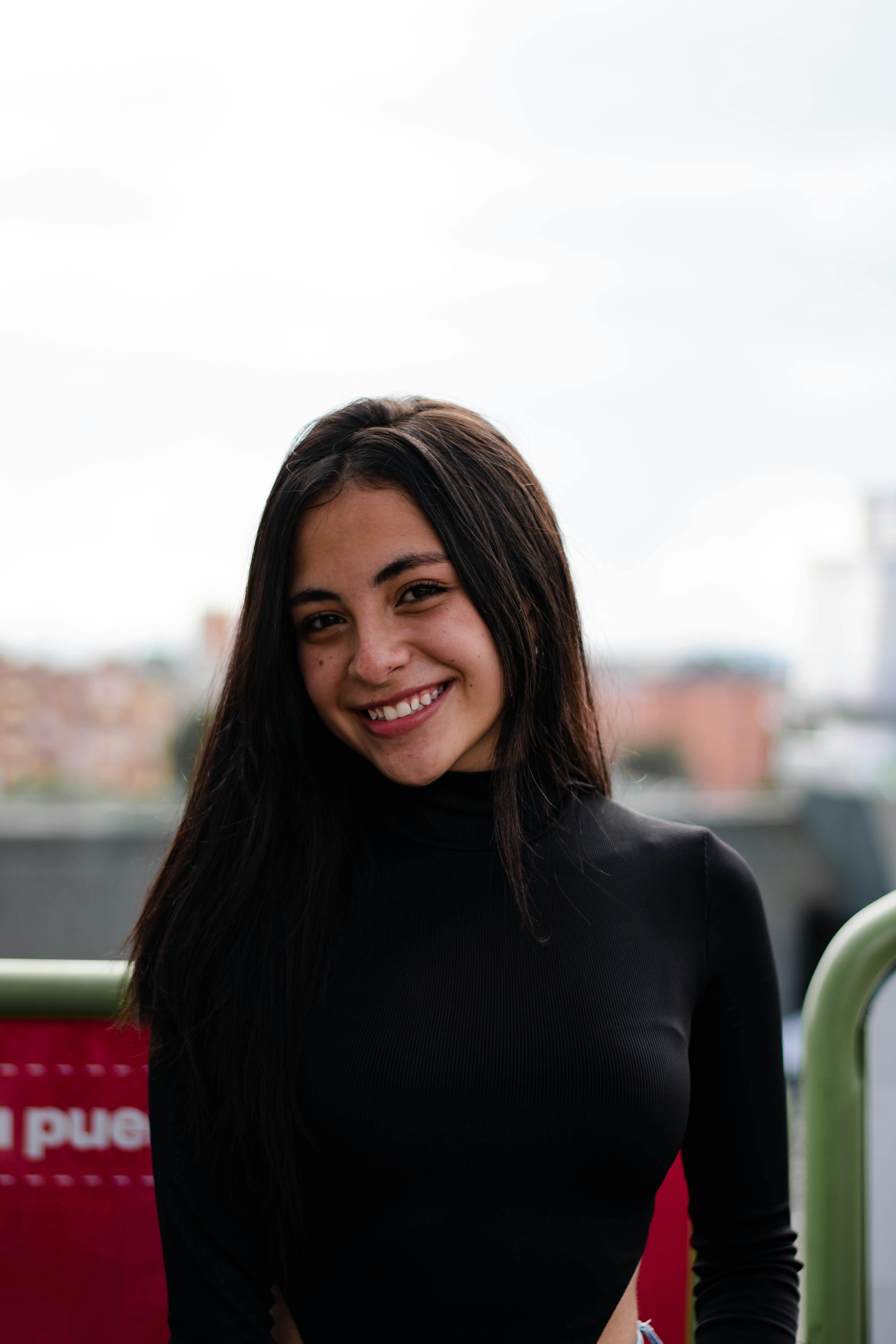 A young woman in a black turtleneck smiles warmly, captured outdoors during the day.