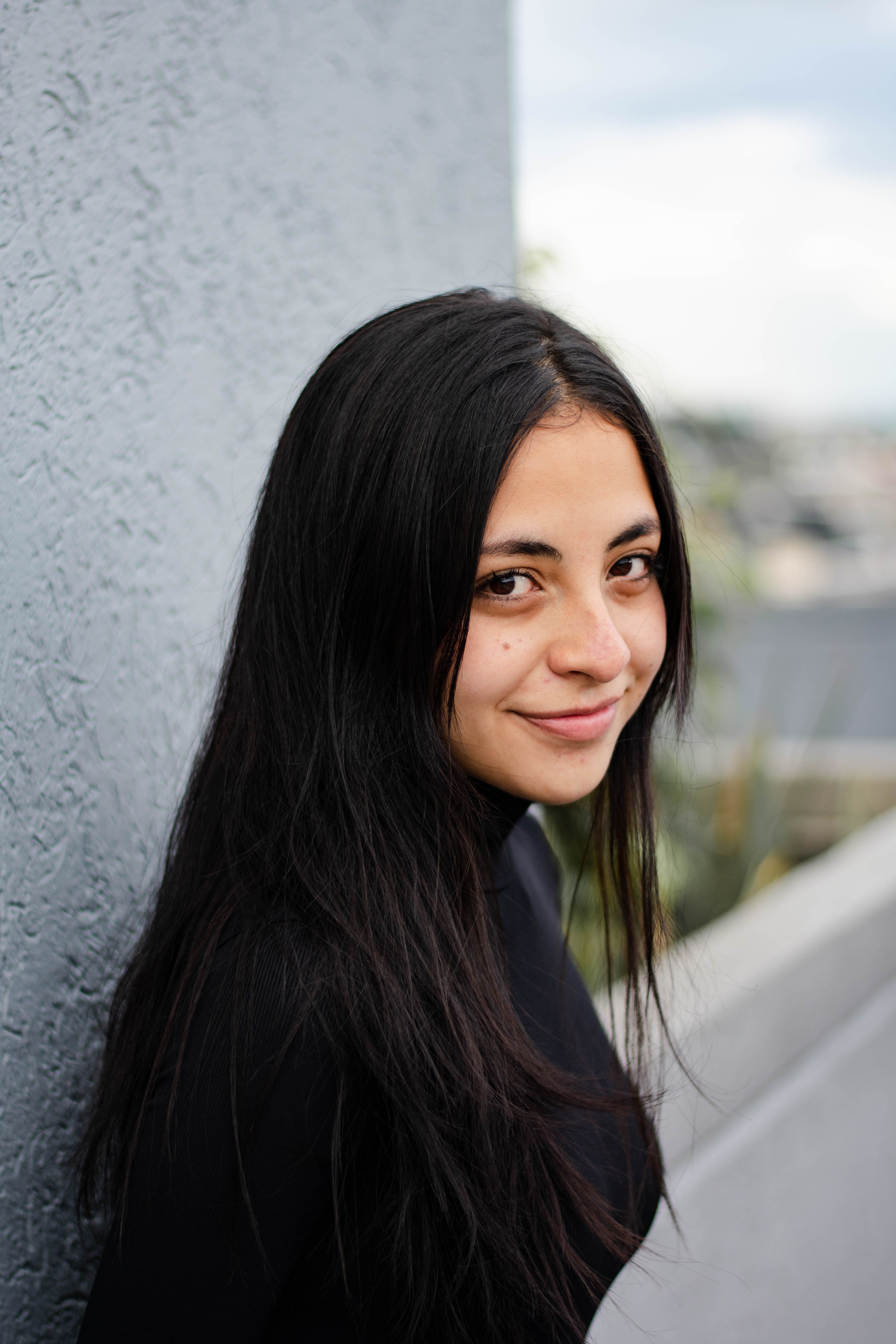 Woman in Black Shirt Leaning on a Gray Wall while Smiling at the Camera