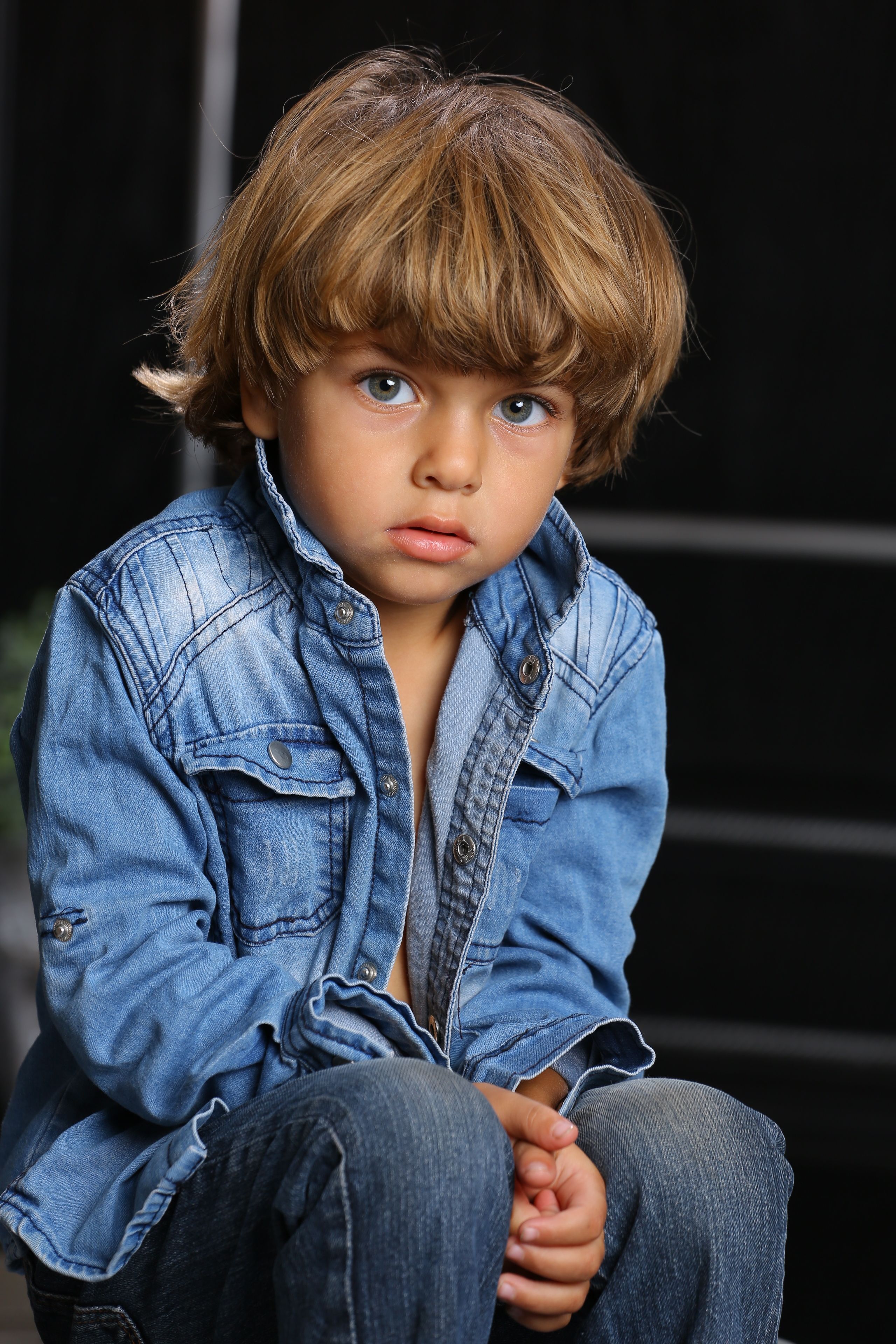 Portrait of a young boy wearing a denim jacket, sitting outdoors with a thoughtful expression.