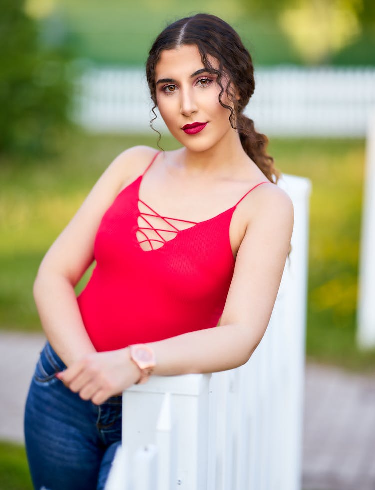 Pretty Young Woman In Red Top And Blue Jeans Leaning Against Fencing