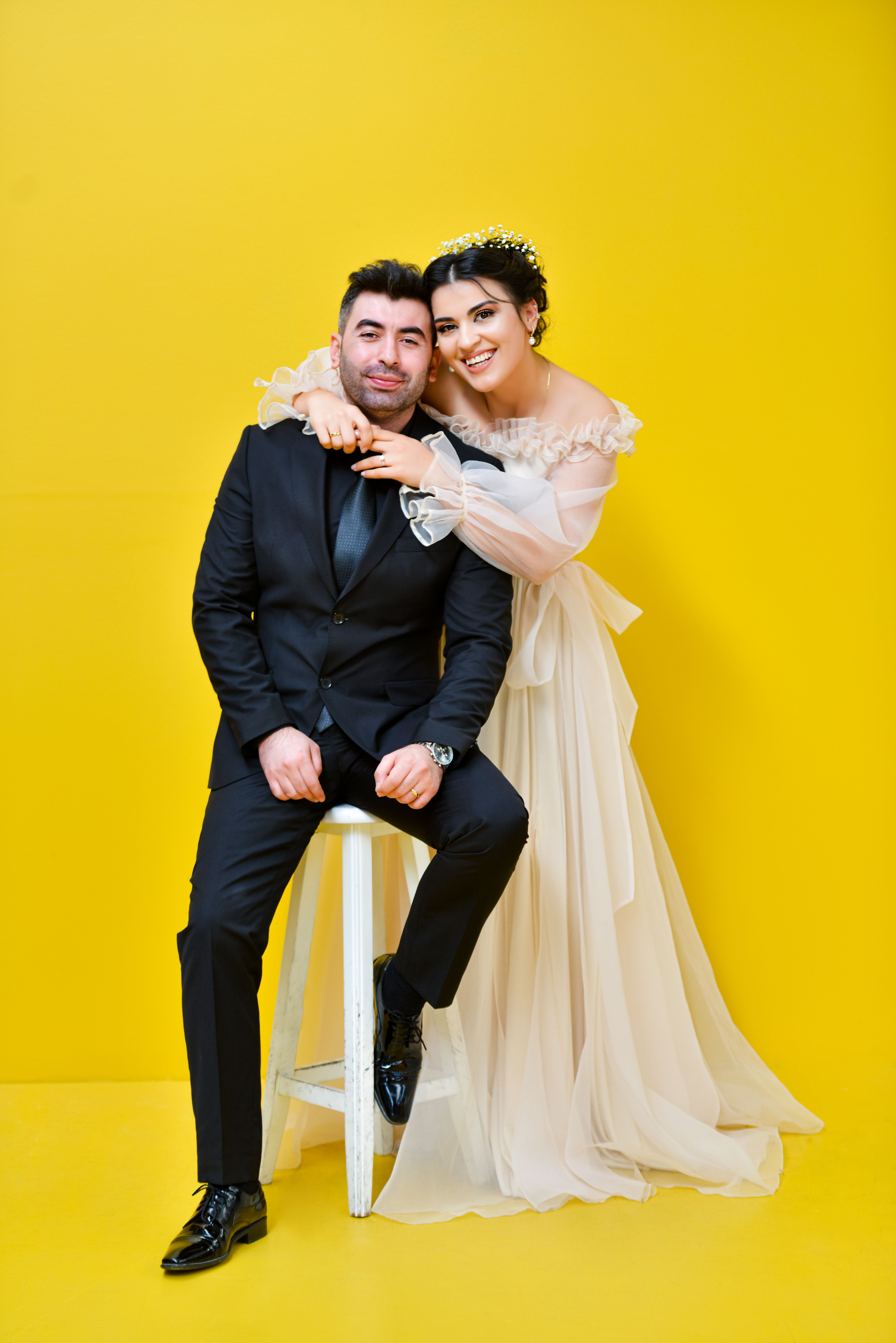 A joyful couple in wedding attire posing against a vibrant yellow background, showcasing love and elegance.