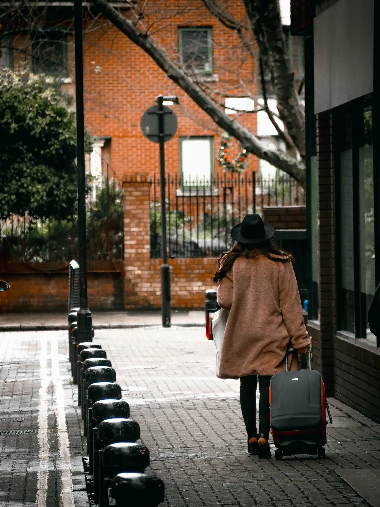tan-coat-hat-fall-travel-outfit Woman in a tan coat and black hat walking with a suitcase in London