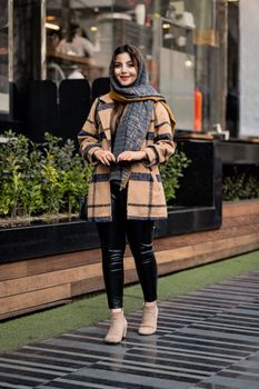 A stylish woman wearing a hijab and plaid coat, smiling on a sidewalk in Tehran.