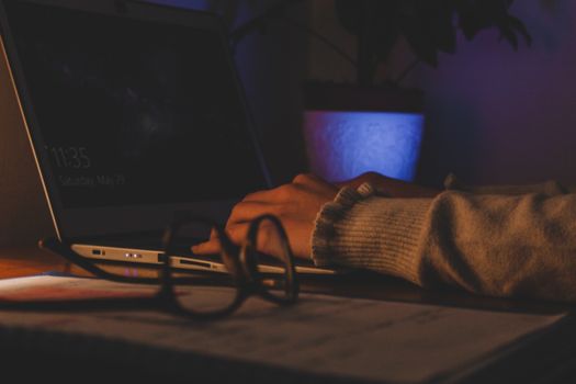 Hands typing on a laptop in a dimly lit room, with eyeglasses in the foreground, creating a cozy atmosphere.