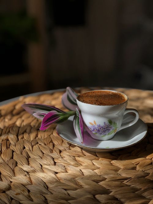 Free A delicate floral porcelain cup filled with rich coffee on a woven placemat, accented by purple flowers. Stock Photo